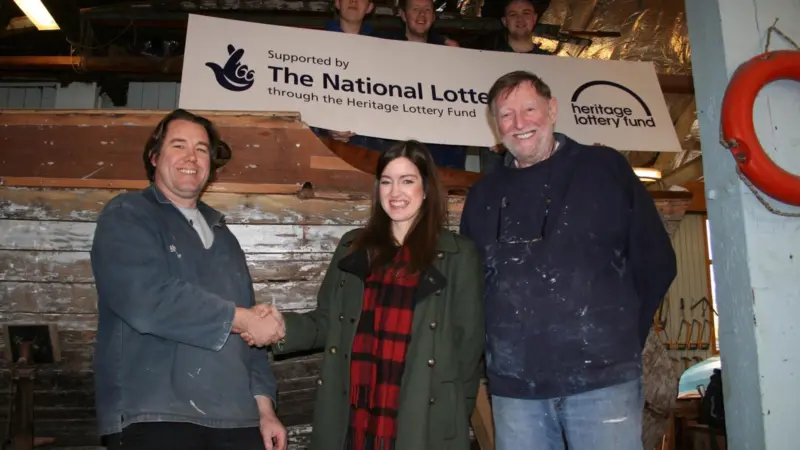 Jonny Bell (heritage boat builder with BTS) celebrates with Ellen Creighton (HLF) and volunteer Phil Charlton as trainees of the Blyth Tall Ship Project look on