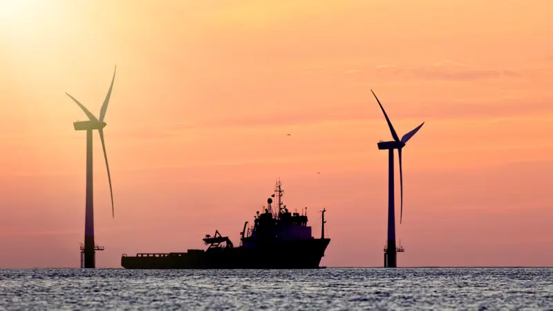 The image shows a vessel at sunset with wind turbines in the background